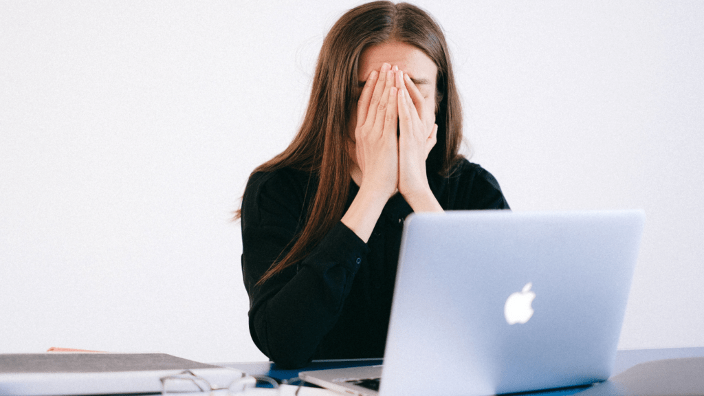 a person covering their face with their hands while sitting at a desk with a laptop