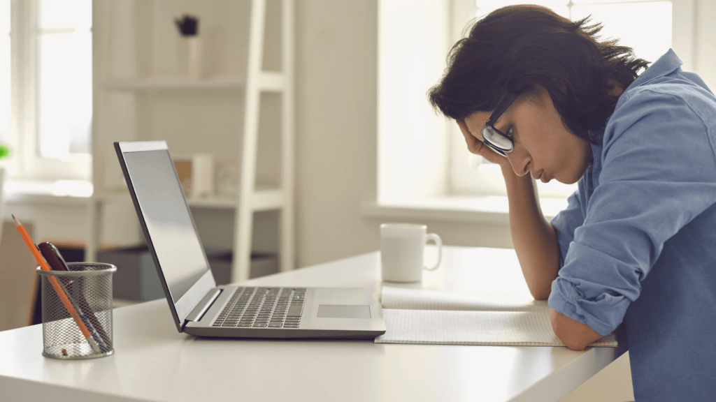 a person covering their face with their hands while sitting at a desk with a laptop